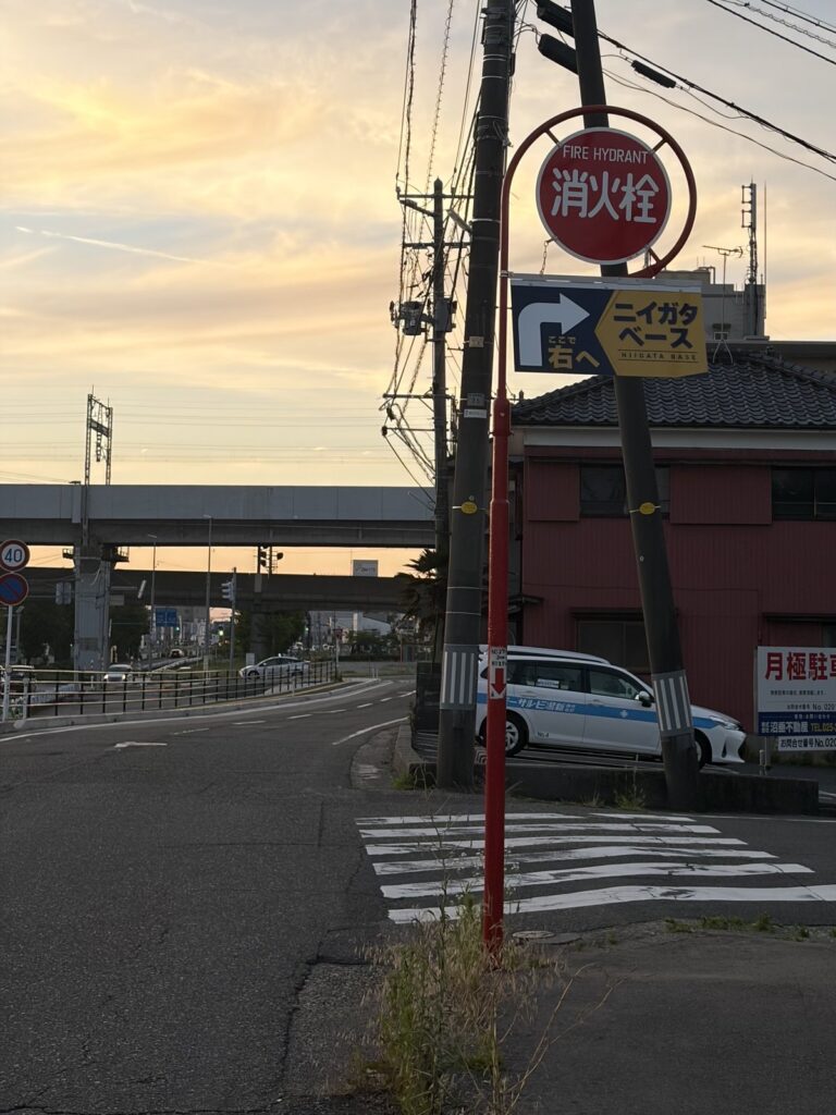交差点の角に立つ赤い消火栓の標識と、「ニイガタベース ここで右へ」の道案内サイン。