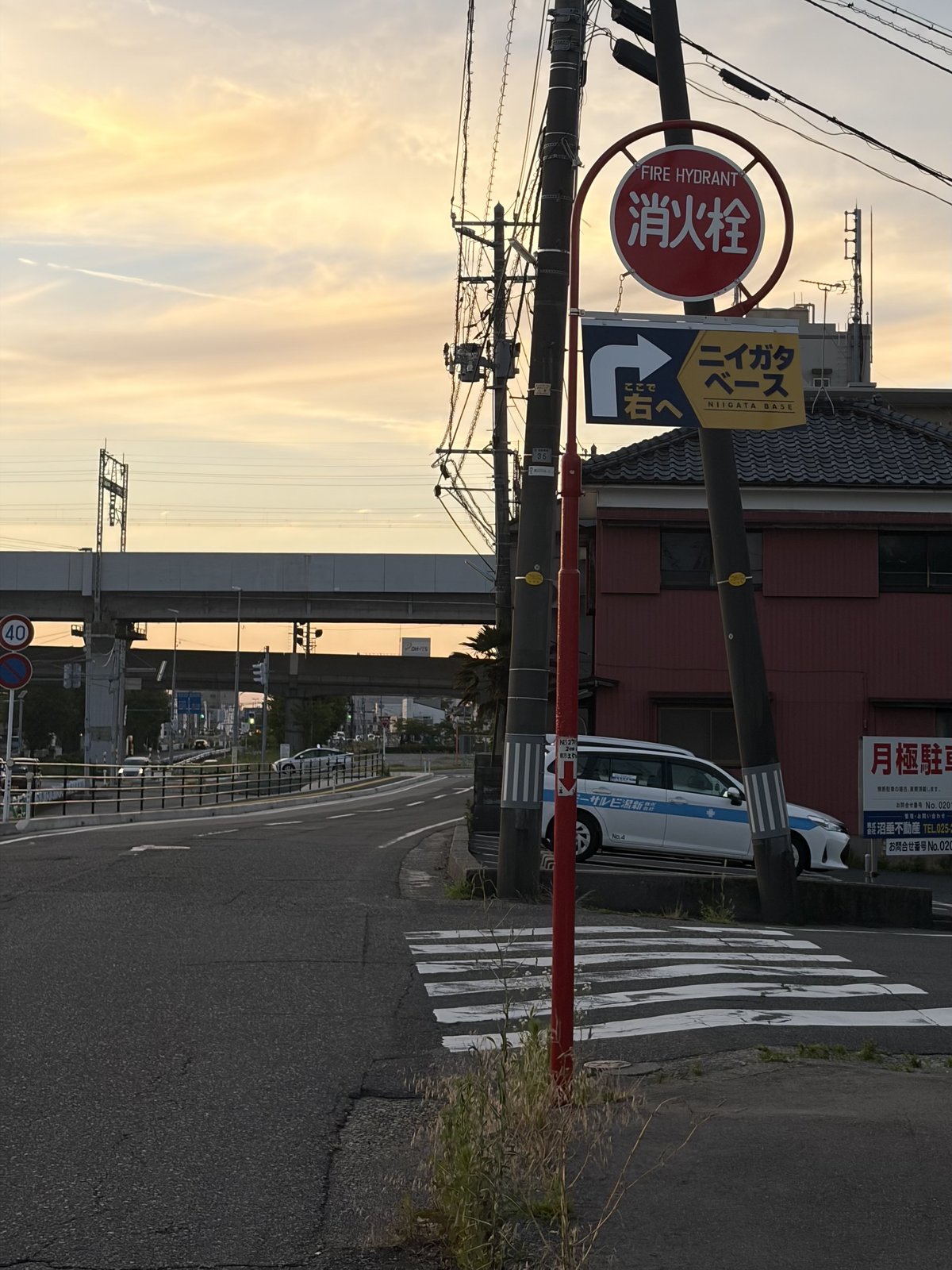 県道5号沿いの交差点の角に立つ赤い消火栓の標識と「ニイガタベース →右へ」の道案内サイン。奥に高架が見える街角の様子。