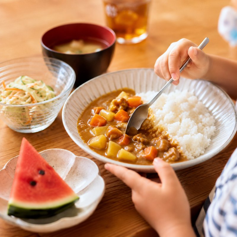 子ども食堂のイメージ。カレーライスとお味噌汁、すいかが並ぶ食卓で、子どもがスプーンを持っている。