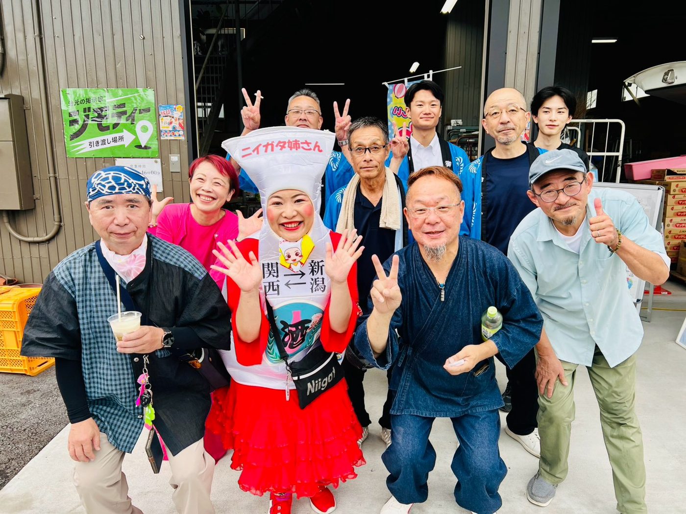 会場の外で、ニイガタ姉さんを囲んで参加者たちが手を振って笑顔でポーズをとっている集合写真。