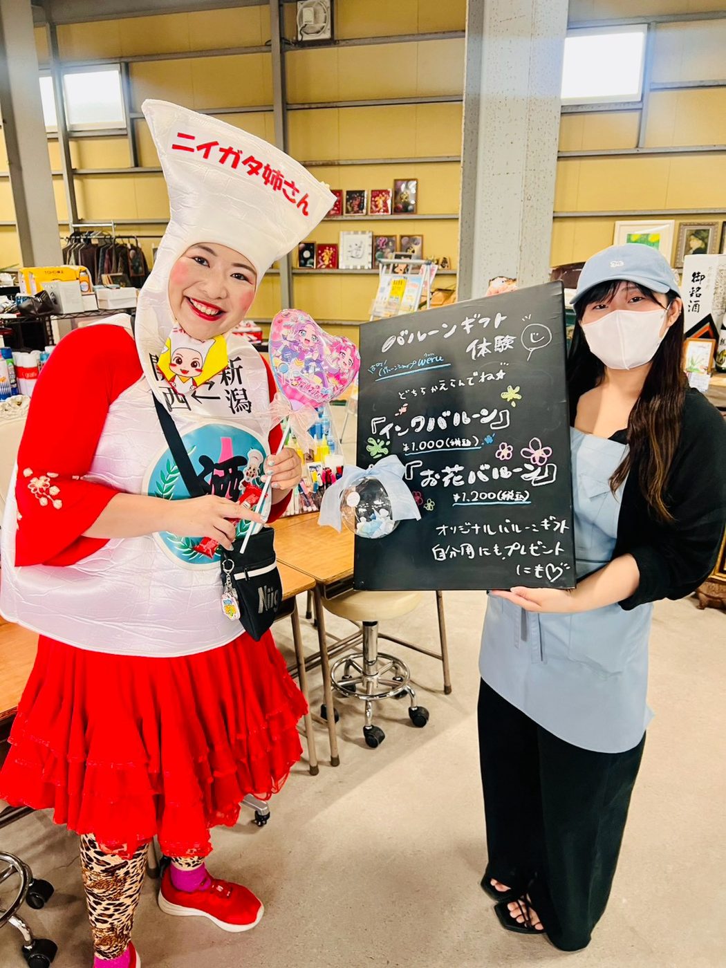 黒板に書かれたバルーンギフト体験の案内の前で、ニイガタ姉さんと出店者が笑顔で立っている。