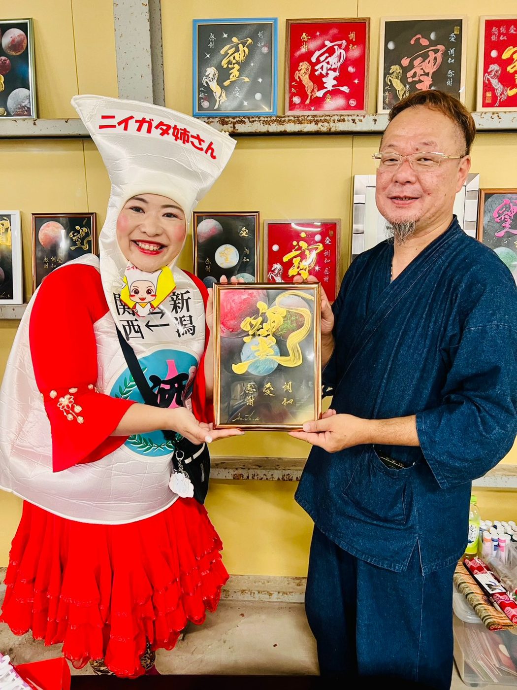 ニイガタ姉さんと甚平姿の男性が、額に入った作品を一緒に持って笑顔でポーズをとっている。