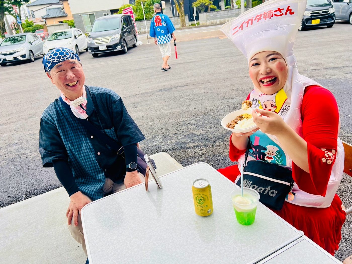 屋外に出したテーブルで、来場者とニイガタ姉さんが飲み物を手に笑顔で座っている。