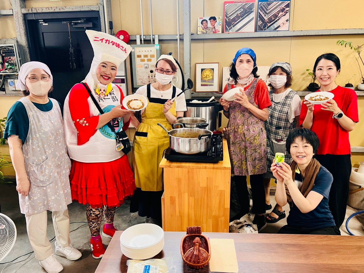 厨房で、エプロン姿のボランティアのみなさんとニイガタ姉さんが、できあがったカレーライスの皿を持って笑顔で並んでいる。