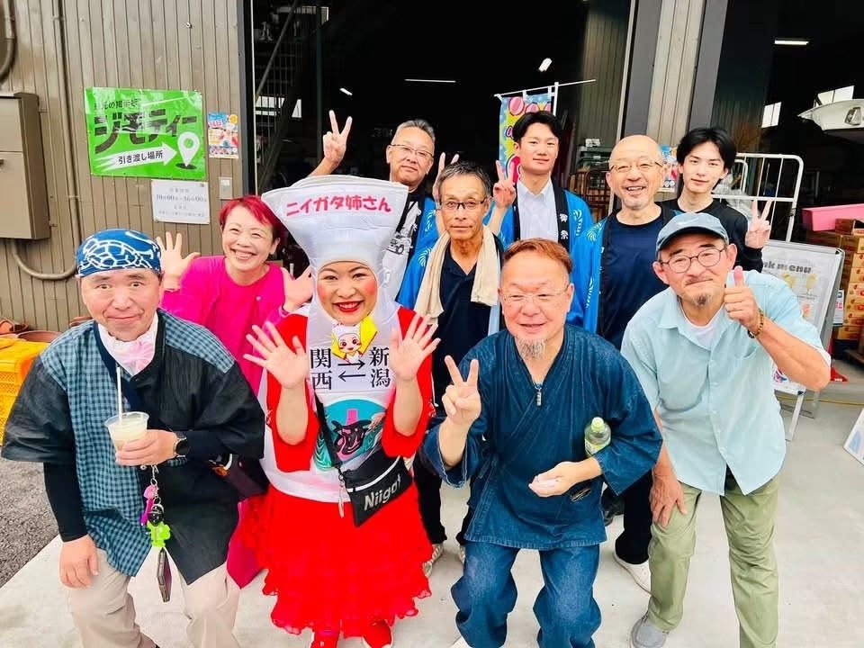 縁日祭の会場入口で、ニイガタ姉さんを囲んで、スタッフや来場者が笑顔でポーズをとっている集合写真。