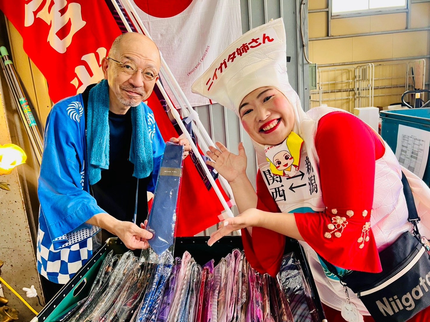 甚平姿の出店者とニイガタ姉さんが、ずらりと並んだネクタイの箱を指さして笑顔で紹介している。