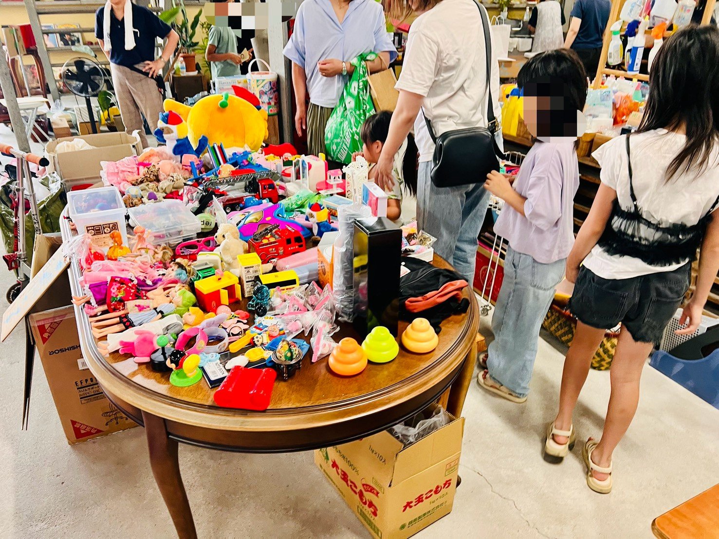 丸いテーブルいっぱいに並んだおもちゃや人形を、子どもたちと保護者が選んでいる様子。
