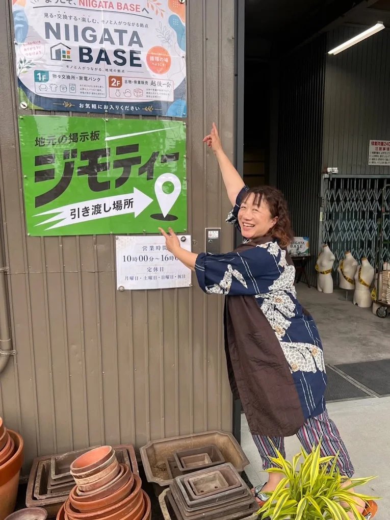 ニイガタベースの入口。NIIGATA BASEの案内板と「地元の掲示板ジモティー 引き渡し場所」のサイン、営業時間の掲示を、浴衣姿のスタッフが笑顔で指さしている。