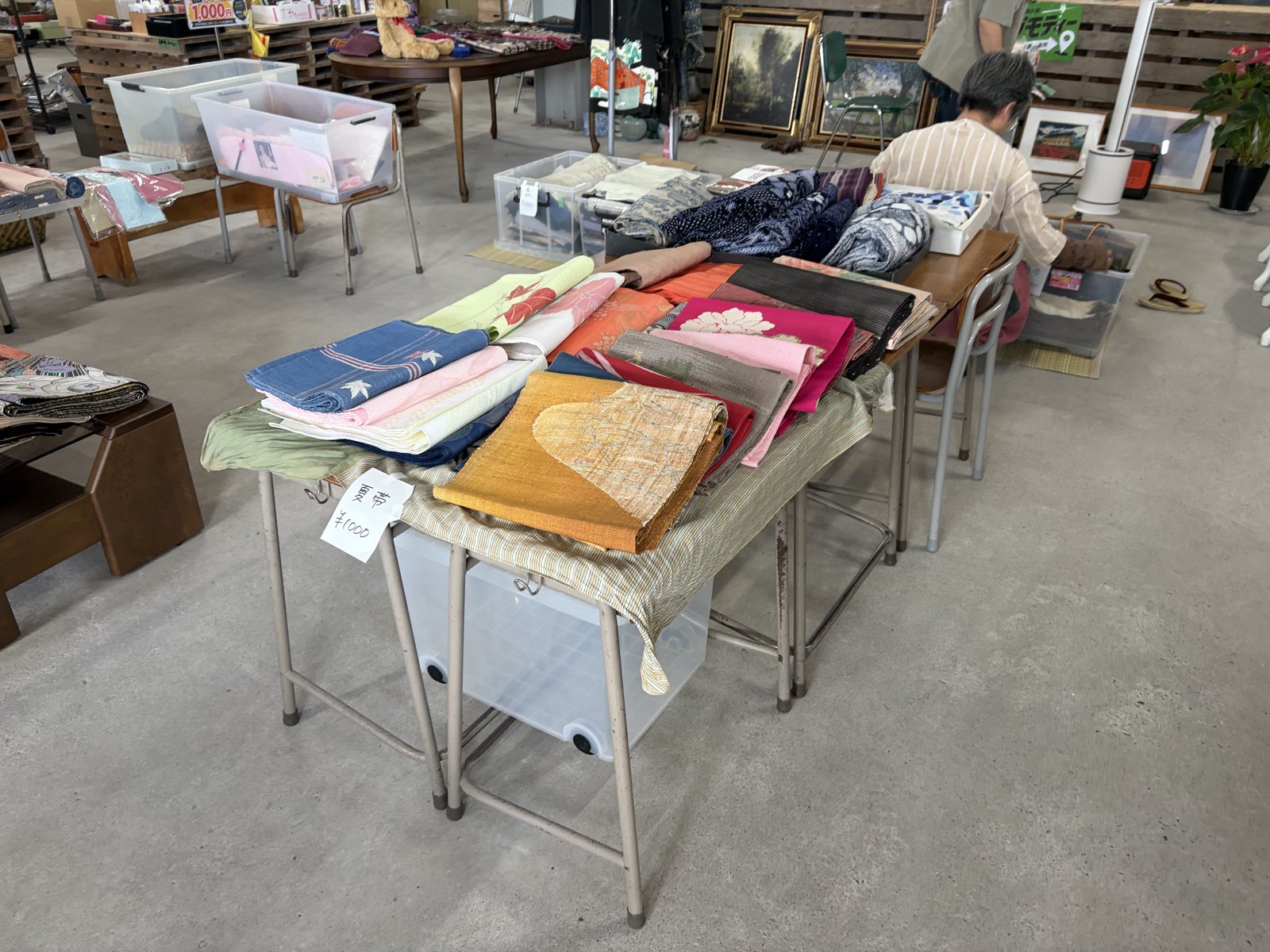 ニイガタベース店内の着物リメイク販売の売り場。テーブルに色とりどりの夏帯や反物が並び、「夏帯 ¥1000」の手書きの値札が添えられている。