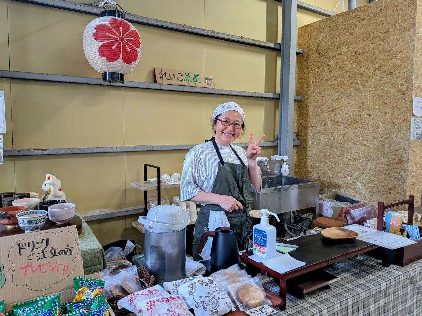 ニイガタベース店内の茶屋コーナー「れいこ茶屋」。桜柄の提灯の下、エプロン姿の店主が笑顔でピースサインをしている。