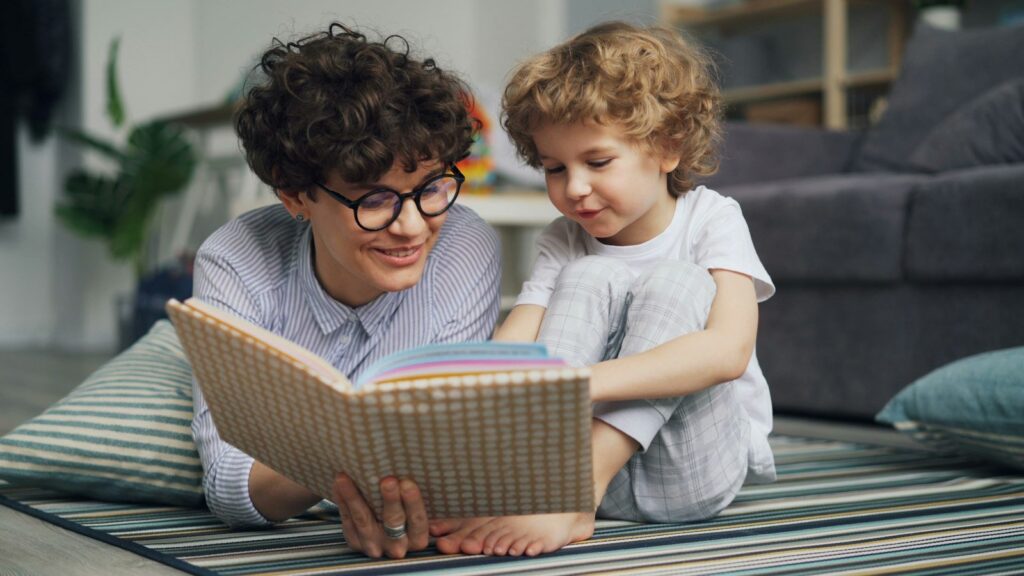 リビングの床で、大人と小さな子どもが一緒に一冊の絵本を広げて読んでいる。