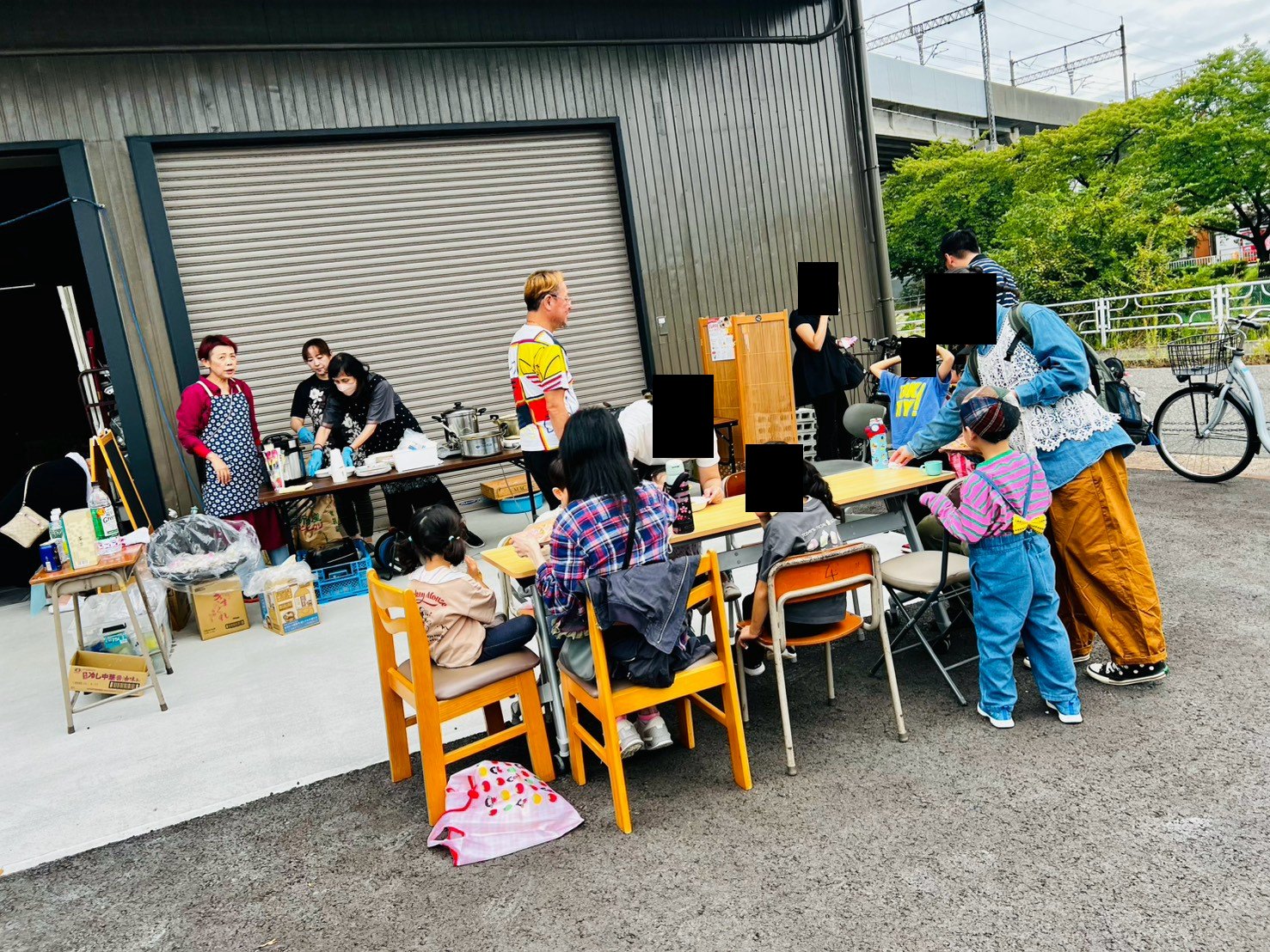 倉庫の前に並べた机を囲んで、子どもや家族連れが座っている。奥では子ども食堂のボランティアが配膳している。来場者の顔はプライバシー保護のため隠している。