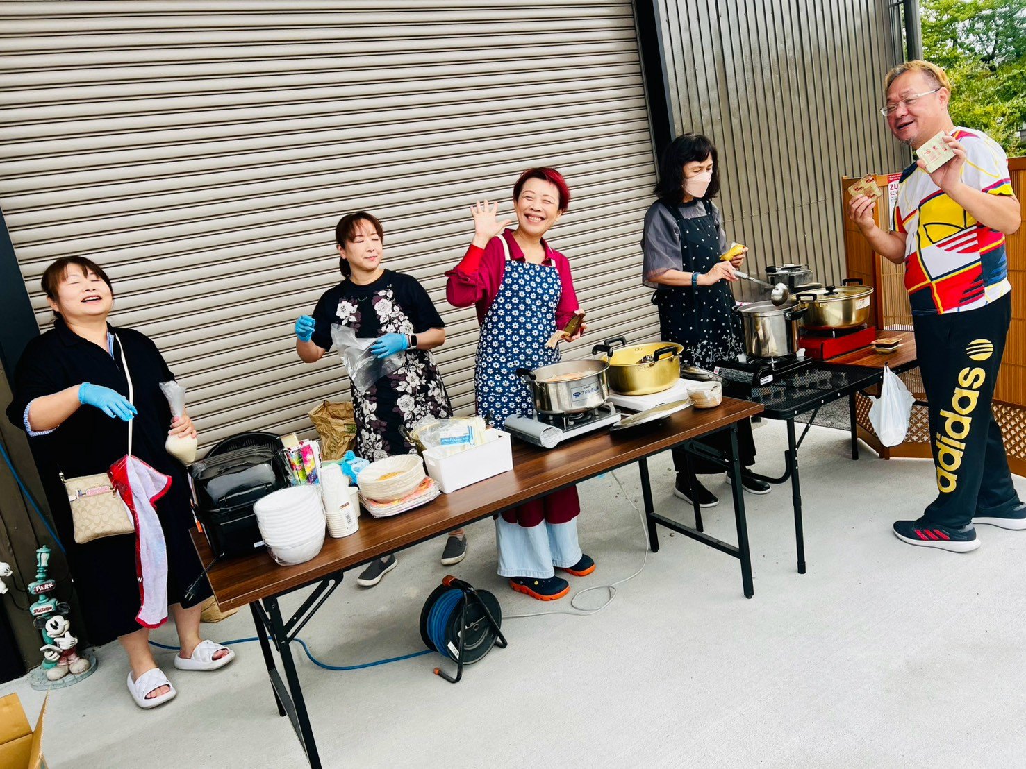 鍋やホットプレートを並べた長机の前で、エプロン姿のボランティアのみなさんが手を振っている。