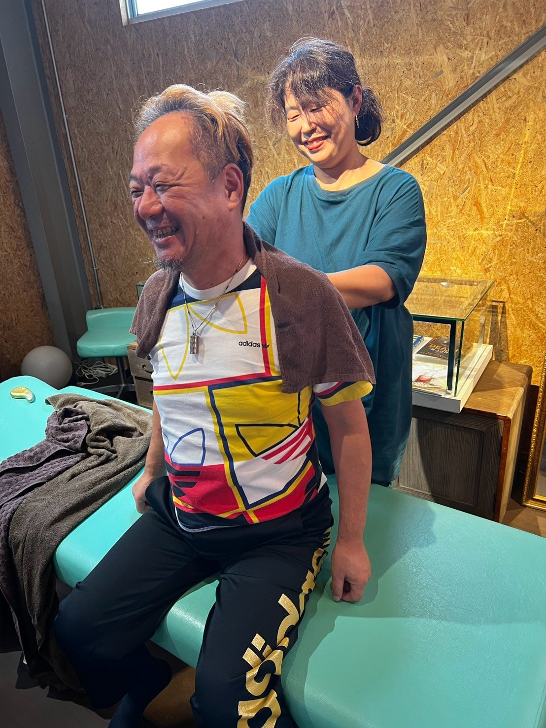 施術台に座った男性の肩を、後ろから女性の整体師がほぐしている。男性は笑っている。