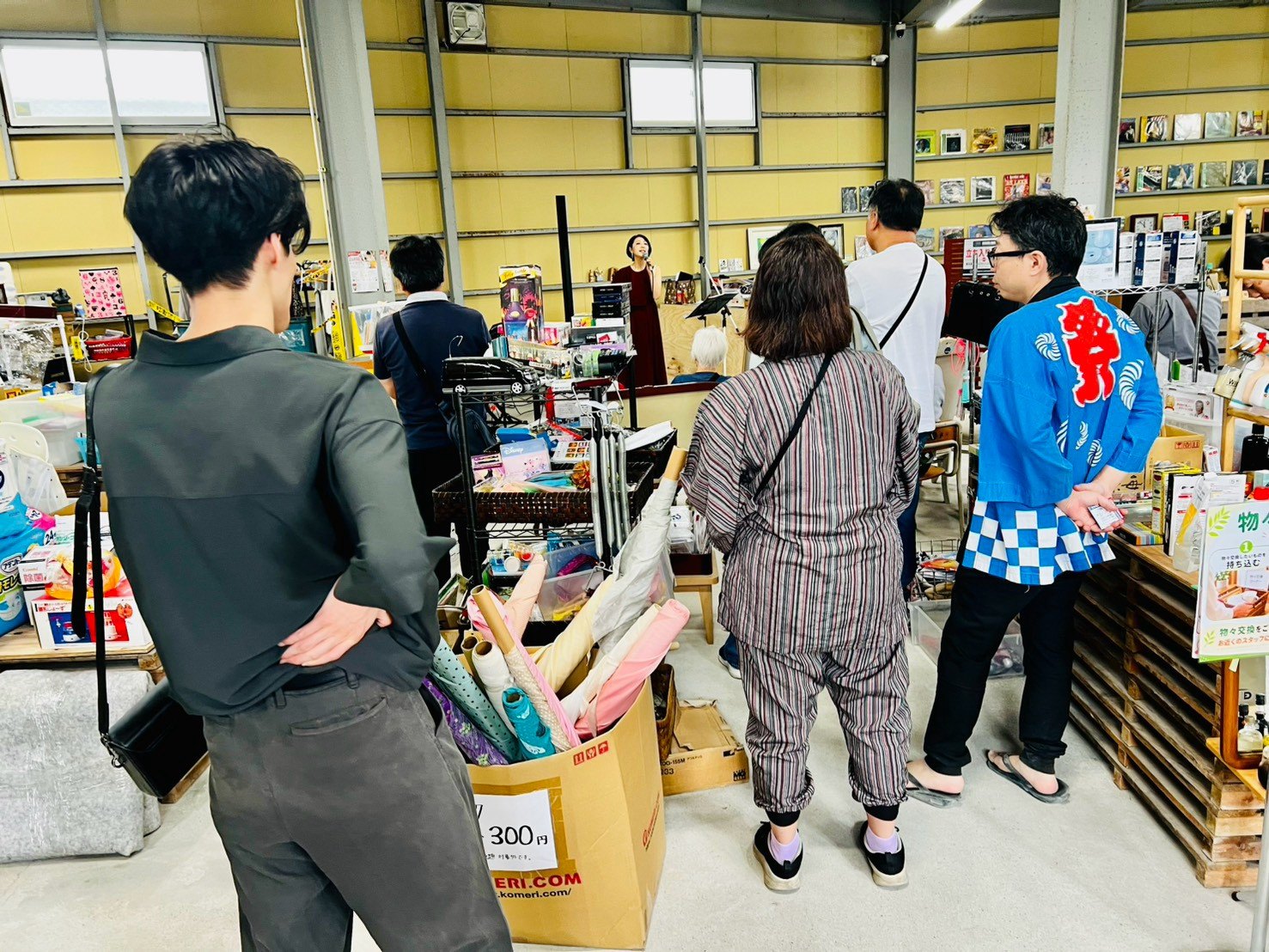 リサイクル品が並ぶ倉庫の中で、来場者が品物を見て回っている後ろ姿。奥では歌唱LIVEが行われている。