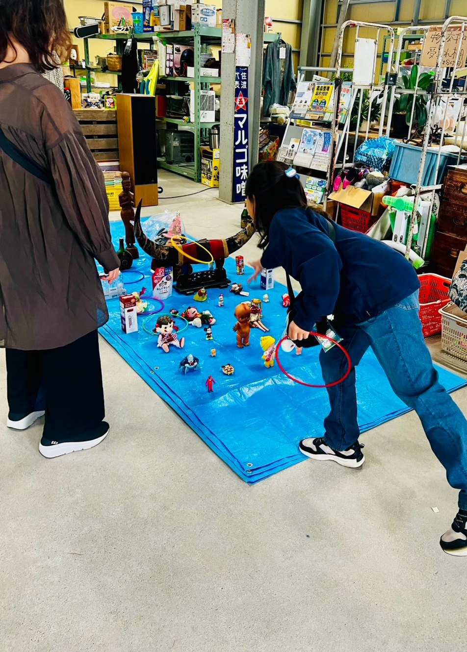 青いシートに並んだ景品に向かって、子どもが身を乗り出して輪を投げようとしている。顔は髪で隠れている。