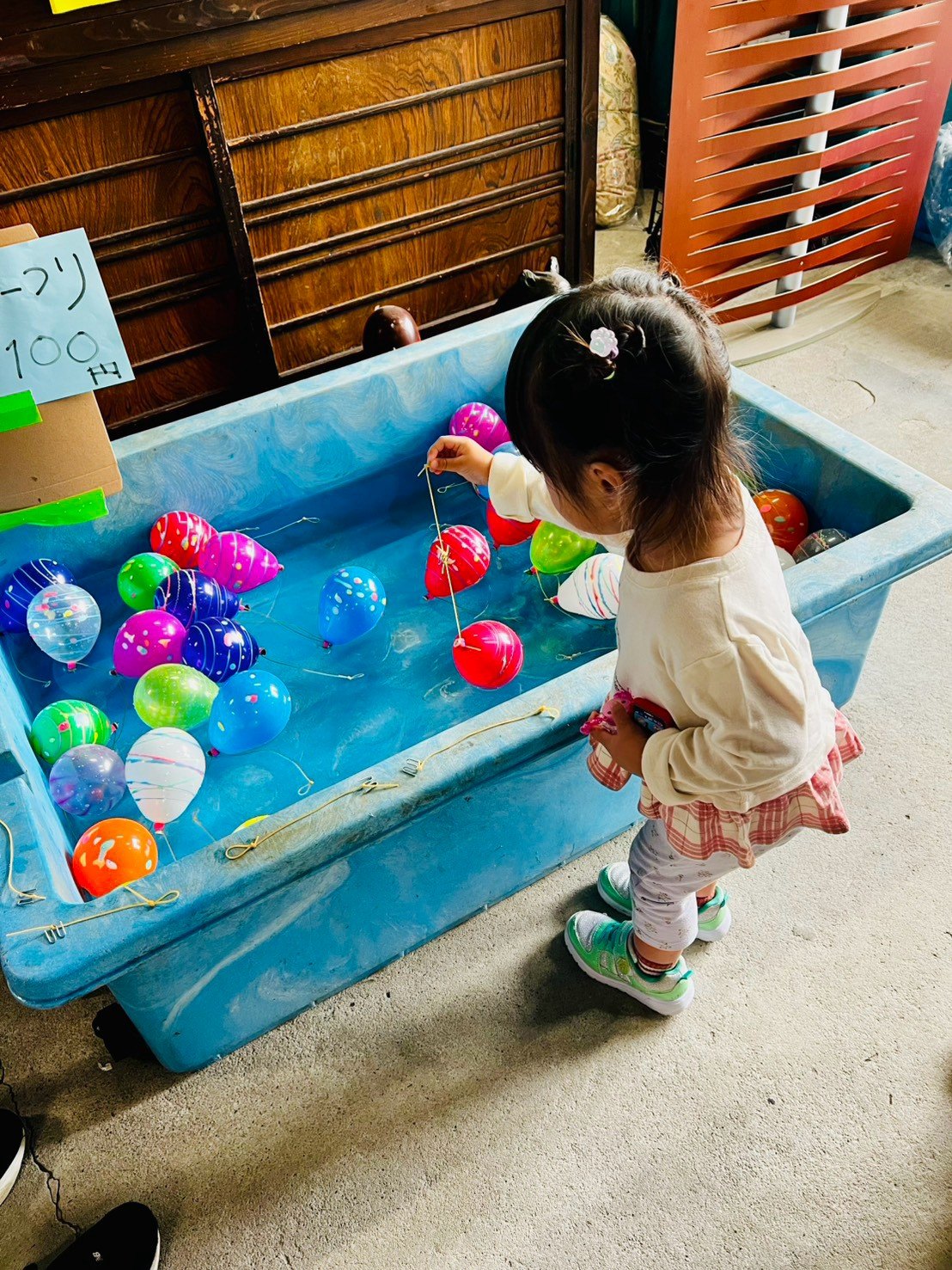 色とりどりのヨーヨーが浮かぶ青い水槽で、小さな女の子がこよりを垂らしてヨーヨーを釣っている後ろ姿。