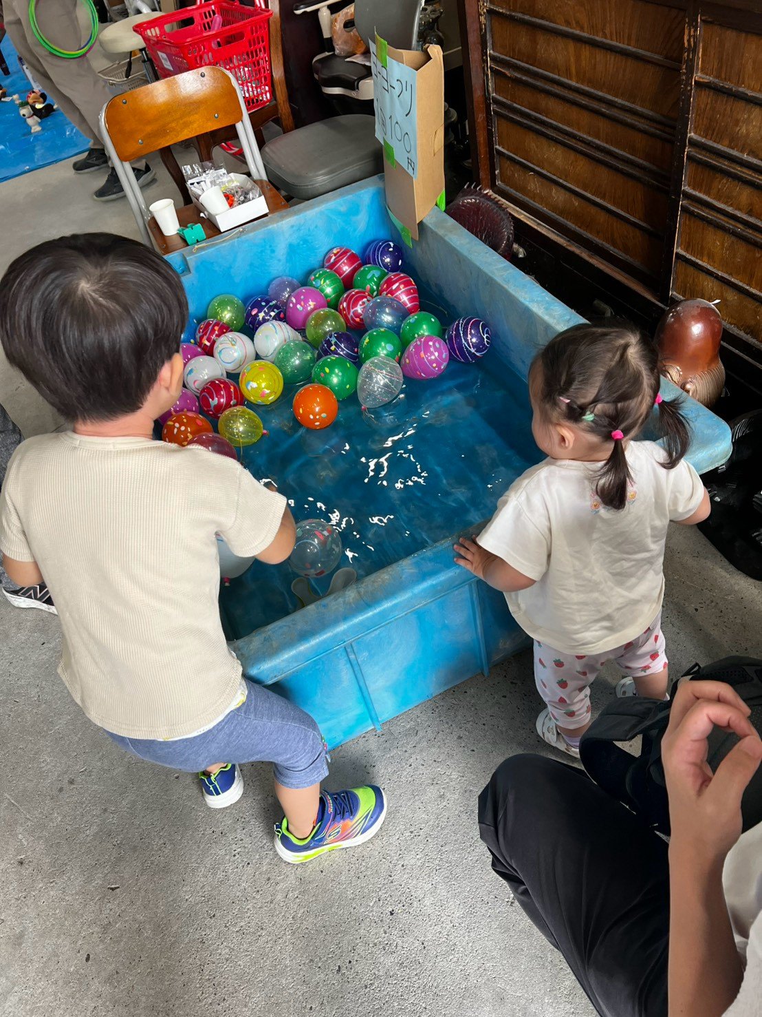色とりどりのヨーヨーが浮かぶ青い水槽を、ふたりの小さな子どもがのぞき込んでいる後ろ姿。
