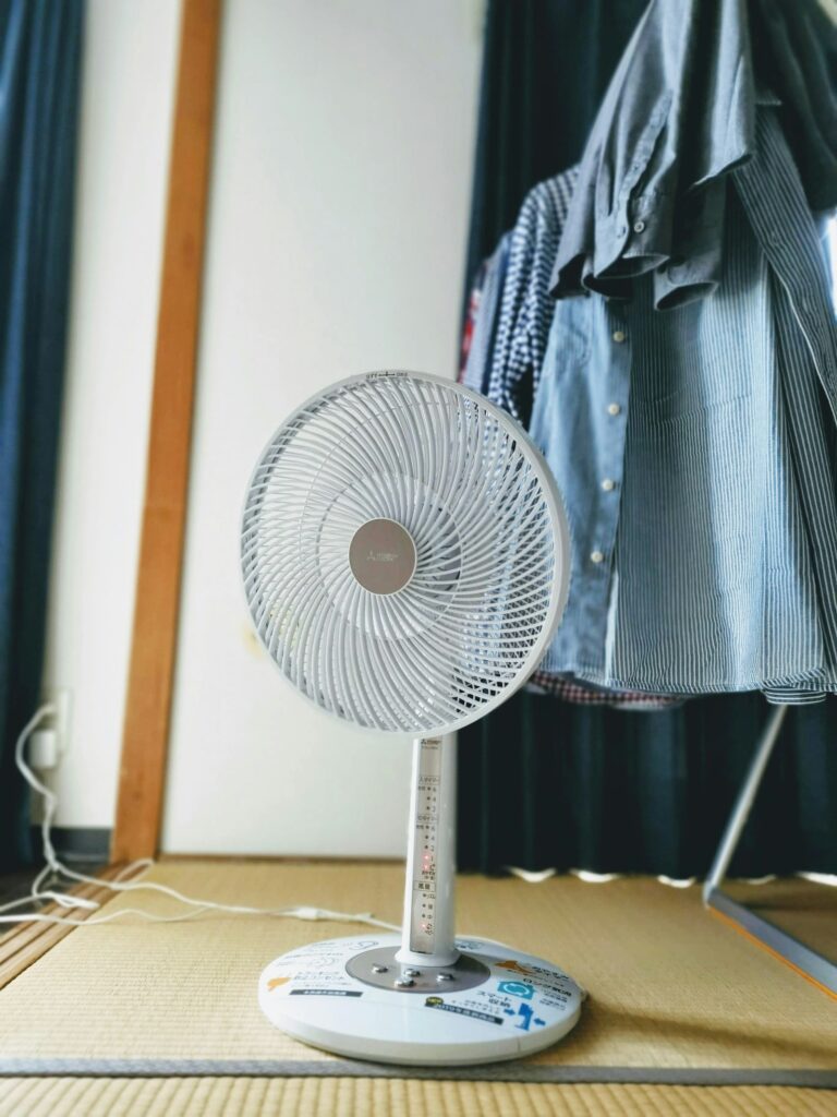 畳の部屋で、ハンガーに掛かったシャツの横に置かれた白い扇風機。
