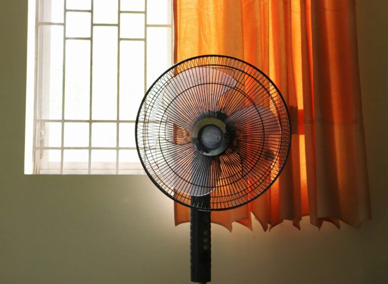 オレンジのカーテンが掛かった窓の前に立つ扇風機。